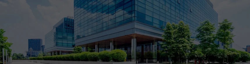 The modern glass office building, home to WorkSuite, boasts reflective windows that mirror the serene trees and bushes around it, all under a partly cloudy sky.