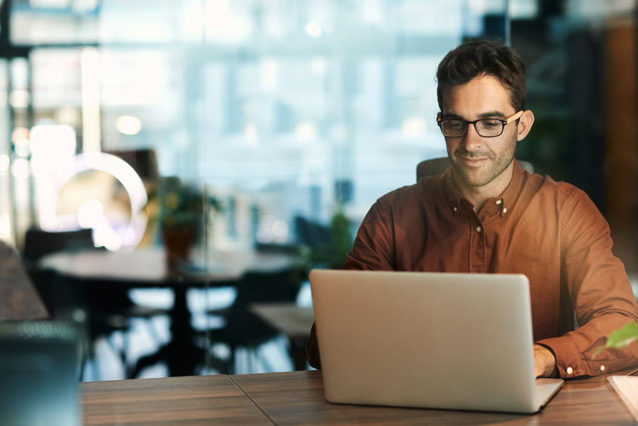 A man wearing glasses and a brown shirt sits at a desk, working on his laptop using MicroStation in a modern office with a blurred background, catering to the needs of 2024 users.