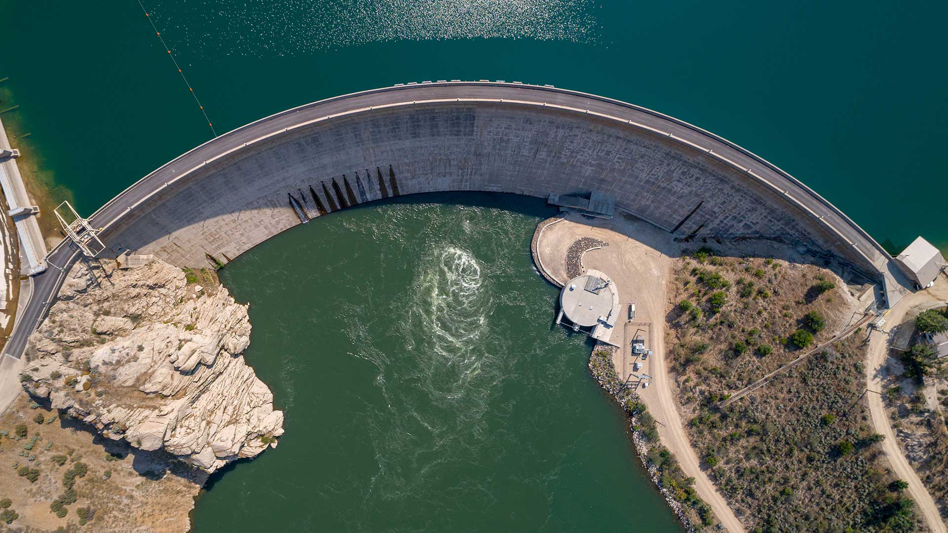 Aerial view of a large dam holding back water, with turbulent water flowing out from the bottom part, surrounded by rugged terrain and a paved road, reminiscent of technical diagrams often found in a CMR Dam Report.