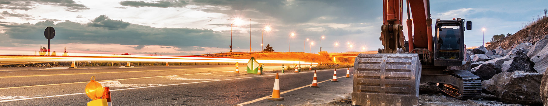 Long exposure image of a road construction site at dusk, featuring streaks of headlight and taillight trails and digital construction equipment to the right.