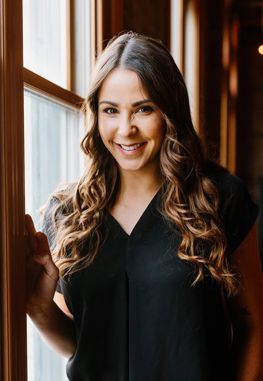 A woman with long brown hair, wearing a black blouse, stands near a window smiling, her laptop open to the latest in OpenRoads Designer roadway design software.