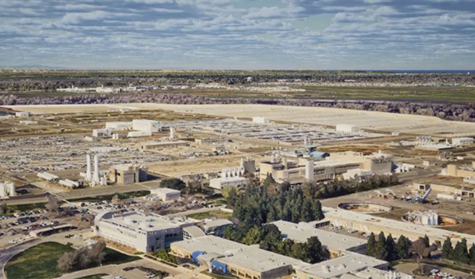 An aerial view of an expansive industrial complex with various buildings and infrastructure. The background features a wide landscape under a partly cloudy sky. The image showcases the integration of photorealistic 3D tiles enabled by iTwin, using Google 3D tiles and Cesium technology.